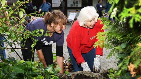 "Eine jüngere und eine ältere Frau mit weißen Haaren gärtnern gemeinsam. Die ältere Frau trägt eine rote Fleecejacke und Gartenhandschuhe. Beide beugen sich konzentriert über ein Pflanzgefäß mit Erde, das im Vordergrund unscharf von grünen Blättern eingerahmt wird."