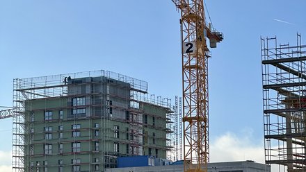 Baustellenaufnahme bei klarem Himmel: großer gelber Turmdrehkran vor mehrgeschossigem Rohbau mit Gerüst, im Vordergrund mehrere orangefarbene Baucontainer.