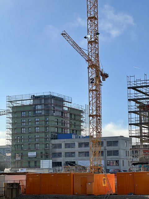 Baustellenaufnahme bei klarem Himmel: großer gelber Turmdrehkran vor mehrgeschossigem Rohbau mit Gerüst, im Vordergrund mehrere orangefarbene Baucontainer.