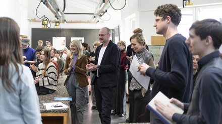 Menschen in einem Atelier/Werkstattraum applaudieren während einer Präsentation; Tische und Hängelampen im Raum. 
