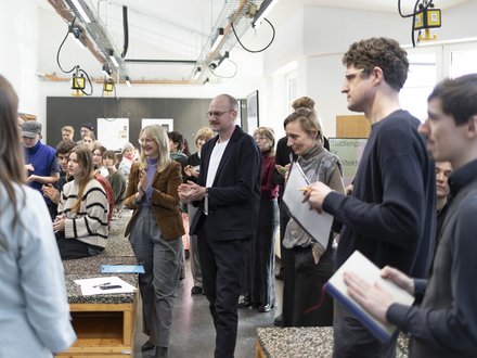 Menschen in einem Atelier/Werkstattraum applaudieren während einer Präsentation; Tische und Hängelampen im Raum. 