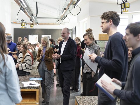 Menschen in einem Atelier/Werkstattraum applaudieren während einer Präsentation; Tische und Hängelampen im Raum. 