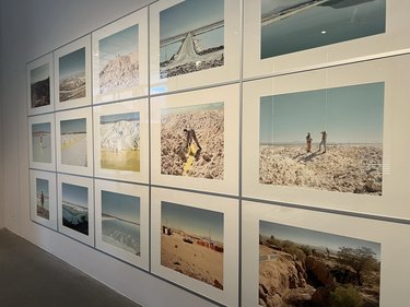 Eine Wand ist vollständig mit gerahmten Fotografien in einem klaren Raster bedeckt. Die Bilder zeigen weite Landschaften: Salzflächen, Wüsten, künstliche Wasserbecken und Menschen bei der Arbeit. Die Farben sind hell, staubig und von Blau- und Beigetönen geprägt. Die Serie dokumentiert Orte des Rohstoffabbaus. Sie macht sichtbar, welche physischen Landschaften und menschlichen Arbeiten hinter digitaler Technologie und Energiespeicherung stehen.