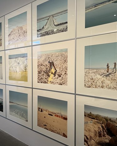 Eine Wand ist vollständig mit gerahmten Fotografien in einem klaren Raster bedeckt. Die Bilder zeigen weite Landschaften: Salzflächen, Wüsten, künstliche Wasserbecken und Menschen bei der Arbeit. Die Farben sind hell, staubig und von Blau- und Beigetönen geprägt. Die Serie dokumentiert Orte des Rohstoffabbaus. Sie macht sichtbar, welche physischen Landschaften und menschlichen Arbeiten hinter digitaler Technologie und Energiespeicherung stehen.