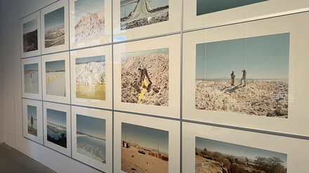 Eine Wand ist vollständig mit gerahmten Fotografien in einem klaren Raster bedeckt. Die Bilder zeigen weite Landschaften: Salzflächen, Wüsten, künstliche Wasserbecken und Menschen bei der Arbeit. Die Farben sind hell, staubig und von Blau- und Beigetönen geprägt. Die Serie dokumentiert Orte des Rohstoffabbaus. Sie macht sichtbar, welche physischen Landschaften und menschlichen Arbeiten hinter digitaler Technologie und Energiespeicherung stehen.