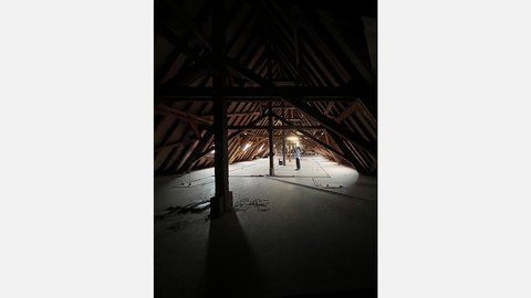 Großer, dunkler Dachboden mit sichtbarem Holztragwerk; einzelne Person steht im beleuchteten Zentrum.