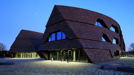 Moderne Schulgebäude mit geneigten Fassaden aus dunklen Holzschindeln auf offenem Platz.