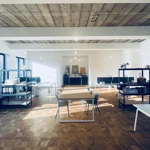 "Heller Großraum-Arbeitsbereich mit Betondecke und weißen Wänden. In der Mitte ein großer Holztisch mit grauen Stühlen. Links und rechts Arbeitsstationen mit dunkelblauen Regalen und Schränken. Im Hintergrund eine Nische mit ausgestellten Kunstwerken. Der Parkettboden zeigt ein geometrisches Muster in Brauntönen. Natürliches Licht fällt durch die linke Fensterfront."