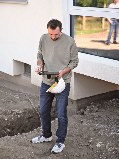 "Ein Mann mit braunem Haar und Bart steht vor einer modernen weißen Hausfassade und hält ein schwarzes Tablet in beiden Händen. Er trägt einen hellgraunen Pullover und dunkelblaue Jeans mit weißen Sneakern. Der Blick ist nach unten auf das Tablet gerichtet. Im Hintergrund ist eine beige Fassade mit großzügigen Fenstern sichtbar, daneben ein Bauplatz mit Erdreich. Durch das große Fenster dahinter sind weitere Personen auf dem Baugelände zu sehen. Eine weiße Rolllade ist oben links im Bild erkennbar."