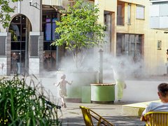 „Ein Kind läuft an einem sonnigen Tag durch den kühlenden Sprühnebel einer urbanen Wasserinstallation auf einem Stadtplatz, während im Vordergrund eine Person auf einem Stuhl sitzt.“