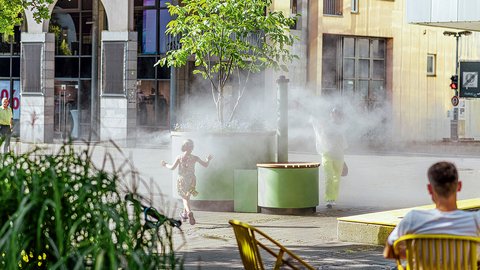 „Ein Kind läuft an einem sonnigen Tag durch den kühlenden Sprühnebel einer urbanen Wasserinstallation auf einem Stadtplatz, während im Vordergrund eine Person auf einem Stuhl sitzt.“