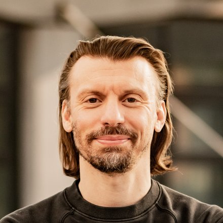 Porträt einer Person mit verschränkten Armen in modernem Gebäude; dunkles Sweatshirt, leichtes Lächeln.