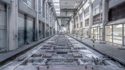 Lange, leere Industriehalle in Zentralperspektive; mittig eine vertiefte, von Metallplatten und Schienen durchzogene Bodenstrecke, rechts ein abgesperrter Gehweg mit Geländern und gelb‑schwarzen Pollern, links große Lüftungskanäle. Oberlichter beleuchten den grauen Raum; keine Personen.