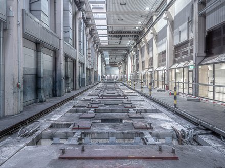 Lange, leere Industriehalle in Zentralperspektive; mittig eine vertiefte, von Metallplatten und Schienen durchzogene Bodenstrecke, rechts ein abgesperrter Gehweg mit Geländern und gelb‑schwarzen Pollern, links große Lüftungskanäle. Oberlichter beleuchten den grauen Raum; keine Personen.