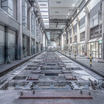 Lange, leere Industriehalle in Zentralperspektive; mittig eine vertiefte, von Metallplatten und Schienen durchzogene Bodenstrecke, rechts ein abgesperrter Gehweg mit Geländern und gelb‑schwarzen Pollern, links große Lüftungskanäle. Oberlichter beleuchten den grauen Raum; keine Personen.