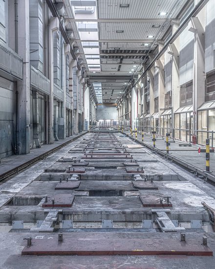 Lange, leere Industriehalle in Zentralperspektive; mittig eine vertiefte, von Metallplatten und Schienen durchzogene Bodenstrecke, rechts ein abgesperrter Gehweg mit Geländern und gelb‑schwarzen Pollern, links große Lüftungskanäle. Oberlichter beleuchten den grauen Raum; keine Personen.
