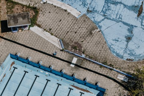 Luftaufnahme eines leerstehenden Freibads mit Becken, Wegen und Treppen.