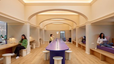 Heller Innenraum mit zentralem Tisch, Sitznischen und Personen beim Arbeiten.