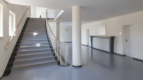 Treppenhalle mit breiter Treppe, Messinghandläufen und runder Stütze; grauer Boden, Türen und Heiznische – schlichte Nachkriegsarchitektur.