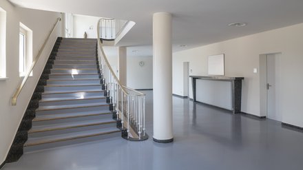 Treppenhalle mit breiter Treppe, Messinghandläufen und runder Stütze; grauer Boden, Türen und Heiznische – schlichte Nachkriegsarchitektur.
