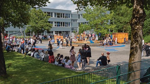 "Ein belebtes Fest auf einem gepflasterten Schulhof bei sonnigem Wetter. Im Hintergrund steht ein mehrstöckiges Schulgebäude. Viele Menschen stehen in Gruppen zusammen, während andere im Schatten am Rand einer Rasenfläche sitzen und in Richtung einer mehrstufigen Holzkonstruktion in der Mitte des Platzes blicken."