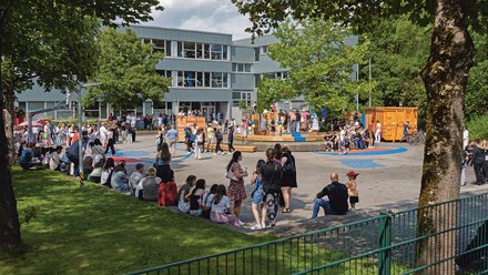 "Ein belebtes Fest auf einem gepflasterten Schulhof bei sonnigem Wetter. Im Hintergrund steht ein mehrstöckiges Schulgebäude. Viele Menschen stehen in Gruppen zusammen, während andere im Schatten am Rand einer Rasenfläche sitzen und in Richtung einer mehrstufigen Holzkonstruktion in der Mitte des Platzes blicken."