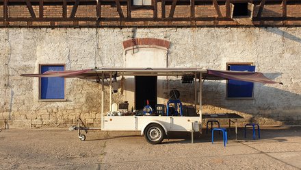 Ein weißer Pkw-Anhänger mit weit aufgeklappten, lilafarbenen Markisenflügeln steht auf einem gepflasterten Hof. Er ist als mobiler Stand mit Arbeitsfläche und Zubehör ausgestattet. Daneben stehen drei blaue Kunststoffhocker an einem kleinen Tisch. Im Hintergrund ist die Fassade eines alten Gebäudes zu sehen: unten weiß verputzt mit blauen Fensterläden, oben ein rustikales Backstein-Fachwerk. Das warme, tiefe Sonnenlicht wirft lange Schatten.