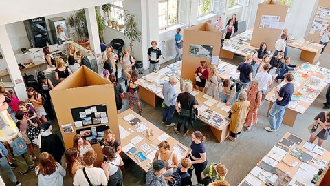 "Vogelperspektive eines hellen Raumes mit vielen Besuchern bei einer Ausstellung oder Veranstaltung. Der Boden ist mit ausgelegten Projekten und Materialien bedeckt. Große aufrecht stehende Pappboxen teilen den Raum in Bereiche. Menschen verschiedenen Alters stehen und gehen zwischen den Ausstellungsbereichen umher. Im Hintergrund sind Fenster, Pflanzen und Büroausstattung sichtbar."