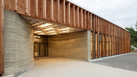 Außenansicht eines eingeschossigen Pavillons mit langer Fassade aus senkrechten Holzlamellen und dahinterliegenden Fensterbändern. Vorn führt eine breite, ebene, stufenlose Fläche unter ein weit auskragendes Vordach in einen überdachten Eingangsbereich. Dort leitet eine geschwungene Stampflehmwand mit sichtbaren Schichtlinien zu einem verglasten Eingang; die Decke darunter ist als helles Raster mit teiltransparenten Lichtfeldern ausgebildet. Warmes Innenlicht fällt nach außen; der Vorplatz besteht aus hellem Beton und Kies, im Hintergrund Bäume unter bewölktem Himmel.