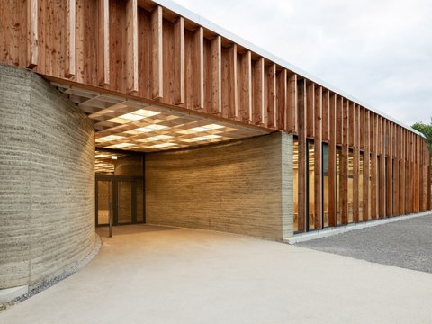 Außenansicht eines eingeschossigen Pavillons mit langer Fassade aus senkrechten Holzlamellen und dahinterliegenden Fensterbändern. Vorn führt eine breite, ebene, stufenlose Fläche unter ein weit auskragendes Vordach in einen überdachten Eingangsbereich. Dort leitet eine geschwungene Stampflehmwand mit sichtbaren Schichtlinien zu einem verglasten Eingang; die Decke darunter ist als helles Raster mit teiltransparenten Lichtfeldern ausgebildet. Warmes Innenlicht fällt nach außen; der Vorplatz besteht aus hellem Beton und Kies, im Hintergrund Bäume unter bewölktem Himmel.