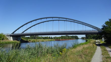Kuhdammbrücke nach Fertigstellung: Schlanke Stahlbogenbrücke über dem Kanal; Uferweg und Vegetation im Vordergrund, klarer Himmel.