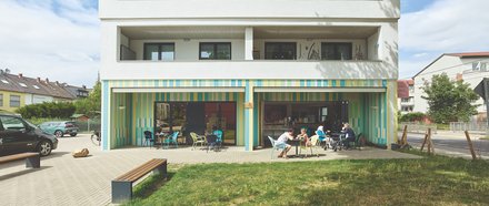 Front eines viergeschossigen, weißen Wohnhauses mit tiefen Loggien; Erdgeschoss als Café mit farbig gestreifter Fassade und offenen Türen; mehrere Menschen sitzen an Tischen im Freien, davor Rasen und Bänke.