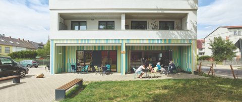 Front eines viergeschossigen, weißen Wohnhauses mit tiefen Loggien; Erdgeschoss als Café mit farbig gestreifter Fassade und offenen Türen; mehrere Menschen sitzen an Tischen im Freien, davor Rasen und Bänke.