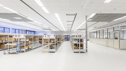 Helle, sehr aufgeräumte Lagerhalle mit weißen Böden und Decken; Reihen niedriger Metallregale mit Kartons, mit B–D beschriftet. Links blaue Transportwagen, rechts eine verglaste Bürotrennwand. Keine Personen.