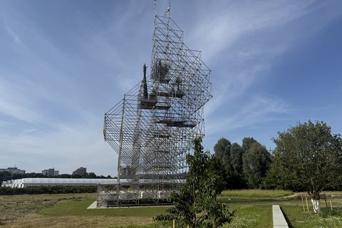  Großflächige Kunstinstallation aus Stahlgerüst auf einer Wiese. Die Gerüstkonstruktion formt eine abstrakte Kopfsilhouette in der Seitenansicht. Im Hintergrund sind Bäume, eine weiße Lagerhalle und Hochhäuser einer Stadt zu sehen. Ein einzelner Baum wächst vor der Installation. Der Himmel ist blau mit wenigen Wolken.