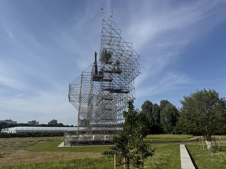  Großflächige Kunstinstallation aus Stahlgerüst auf einer Wiese. Die Gerüstkonstruktion formt eine abstrakte Kopfsilhouette in der Seitenansicht. Im Hintergrund sind Bäume, eine weiße Lagerhalle und Hochhäuser einer Stadt zu sehen. Ein einzelner Baum wächst vor der Installation. Der Himmel ist blau mit wenigen Wolken.