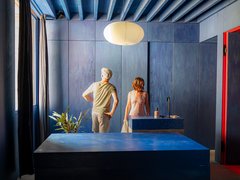 Ein komplett in tiefem Blau gestaltetes Schlafzimmer mit markanter Balkendecke. Zwei Personen stehen im Hintergrund an einem Fenster, davor befindet sich ein massives, blockartiges Möbelstück in der Raummitte.