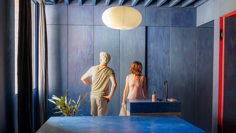 Ein komplett in tiefem Blau gestaltetes Schlafzimmer mit markanter Balkendecke. Zwei Personen stehen im Hintergrund an einem Fenster, davor befindet sich ein massives, blockartiges Möbelstück in der Raummitte.