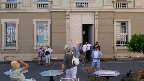 "Außencafé vor einem klassizistischen Gebäude mit Sandsteinfassade. Im Vordergrund sitzen Besucherinnen und Besucher an runden Tischen mit Stühlen. Im Hintergrund steht eine Gruppe von Menschen vor dem Haupteingang mit zwei großen Flügeltüren. Das mehrstöckige Gebäude hat vier große Fenster in der Erdgeschosszone und mehrere kleinere Fenster in der oberen Etage mit Baluster-Brüstungen."