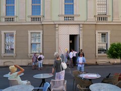 "Außencafé vor einem klassizistischen Gebäude mit Sandsteinfassade. Im Vordergrund sitzen Besucherinnen und Besucher an runden Tischen mit Stühlen. Im Hintergrund steht eine Gruppe von Menschen vor dem Haupteingang mit zwei großen Flügeltüren. Das mehrstöckige Gebäude hat vier große Fenster in der Erdgeschosszone und mehrere kleinere Fenster in der oberen Etage mit Baluster-Brüstungen."