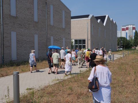 "Große Gruppe von etwa 25 Personen verschiedenen Alters steht auf einem Platz vor modernen Ziegelbau-Gebäuden. Die Menschen tragen leichte Sommerkleidung und einige haben Hüte oder Regenschirme. Im Vordergrund rechts eine ältere Frau in weißem Shirt mit brauner Umhängetasche. Der Himmel ist blau, die Wiese am Rand wirkt trocken und ungepflegt."