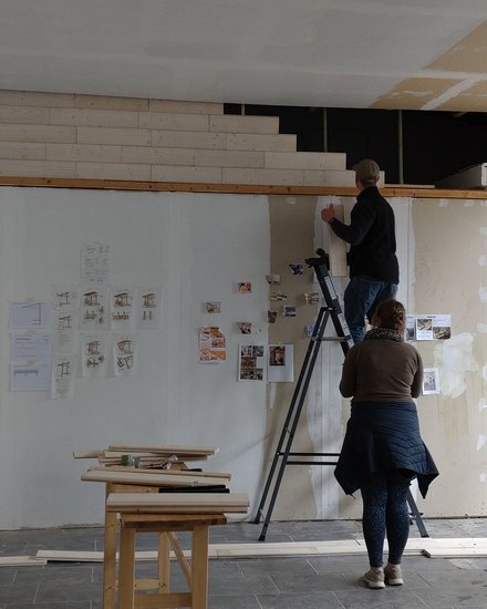 Werkstatt/Studio: zwei Personen, eine auf einer Leiter, befestigen Skizzen und Fotos an einer hohen Wand; Holzleisten, Tische und weitere Leitern stehen im Raum.