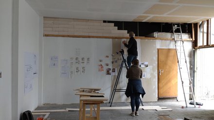 Werkstatt/Studio: zwei Personen, eine auf einer Leiter, befestigen Skizzen und Fotos an einer hohen Wand; Holzleisten, Tische und weitere Leitern stehen im Raum.