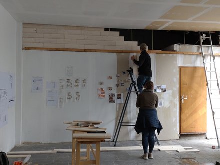 Werkstatt/Studio: zwei Personen, eine auf einer Leiter, befestigen Skizzen und Fotos an einer hohen Wand; Holzleisten, Tische und weitere Leitern stehen im Raum.