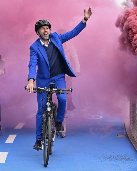Mann im blauen Anzug fährt Fahrrad über die Brücke, farbiger Rauch im Hintergrund.