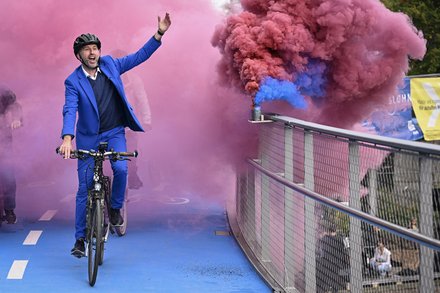 Mann im blauen Anzug fährt Fahrrad über die Brücke, farbiger Rauch im Hintergrund.
