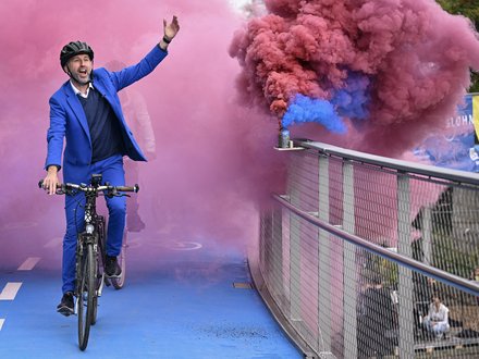 Mann im blauen Anzug fährt Fahrrad über die Brücke, farbiger Rauch im Hintergrund.