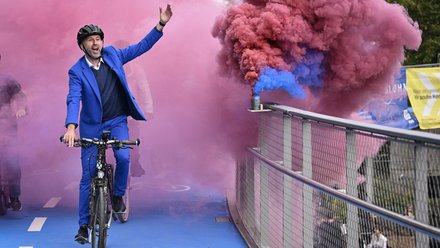 Mann im blauen Anzug fährt Fahrrad über die Brücke, farbiger Rauch im Hintergrund.