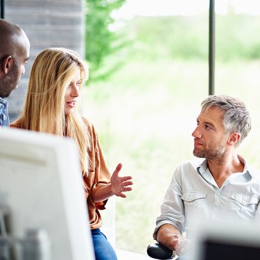 Personen sitzen in einem modernen Büro vor großen Fenstern und unterhalten sich in entspannter Atmosphäre.