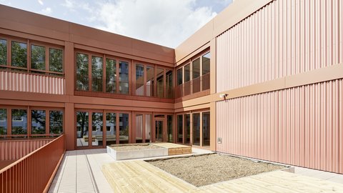 Innenhof eines modernen Schulgebäudes mit Holzterrasse, Sitzbank und großen Fenstern